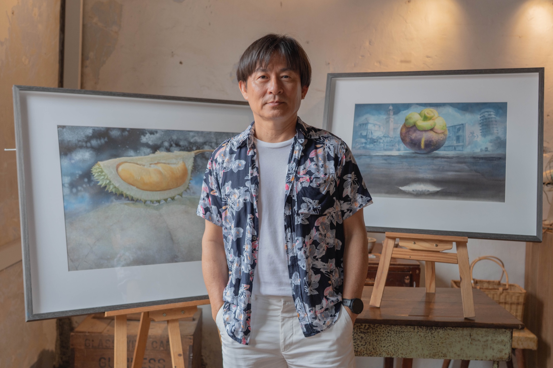 Angelo Chia Seng Chai standing between two framed watercolour paintings of tropical fruits